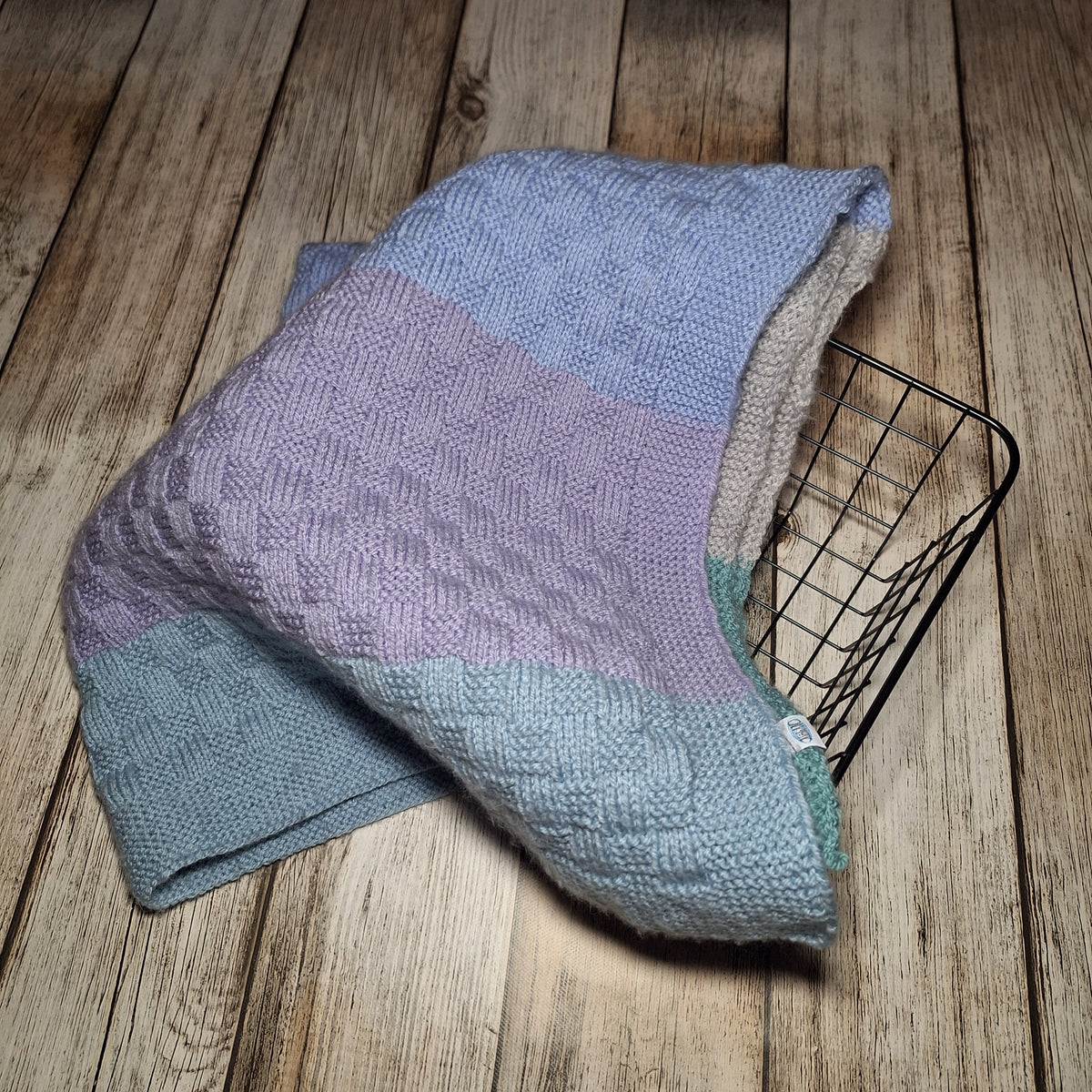Handgestrickte Babydecke mit Pastellstreifen, locker über einen schwarzen Drahtkorb gelegt.
