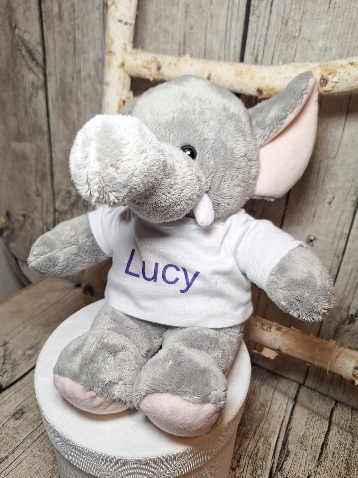 Grauer Plüschelefant mit weißem T-Shirt und dem Namen Lucy in violetter Schrift.