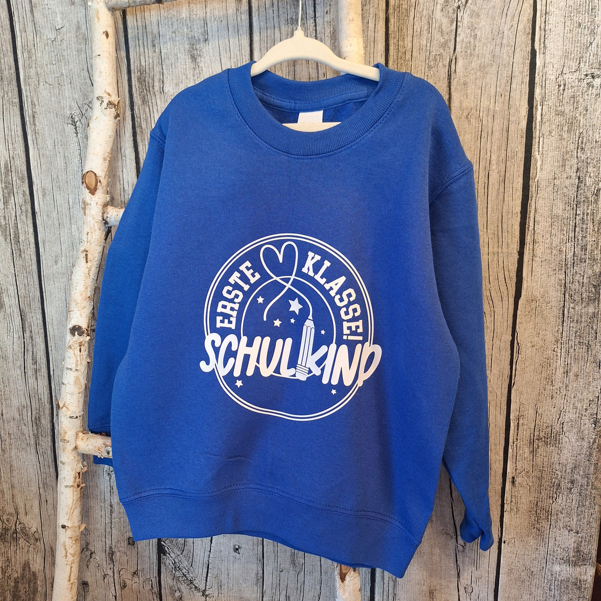 Blauer Kindersweater mit hellem Einschulungsmotiv „Erste Klasse! Schulkind“ auf einem Kleiderbügel vor Holz.