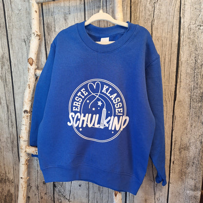 Blauer Kindersweater mit hellem Einschulungsmotiv „Erste Klasse! Schulkind“ auf einem Kleiderbügel vor Holz.