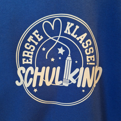 Nahaufnahme des hellen Aufdrucks „Erste Klasse! Schulkind“ mit Bleistift, Herz und Sternen auf blauem Sweatstoff.