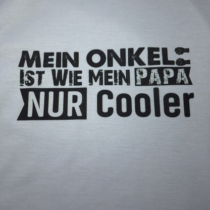 Nahaufnahme des schwarzen Schriftzugs „Mein Onkel ist wie mein Papa – nur cooler“ auf weißem T-Shirt.
