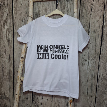 Weißes Kinder-T-Shirt mit schwarzem Spruch „Mein Onkel ist wie mein Papa – nur cooler“ auf einem Holzkleiderbügel.