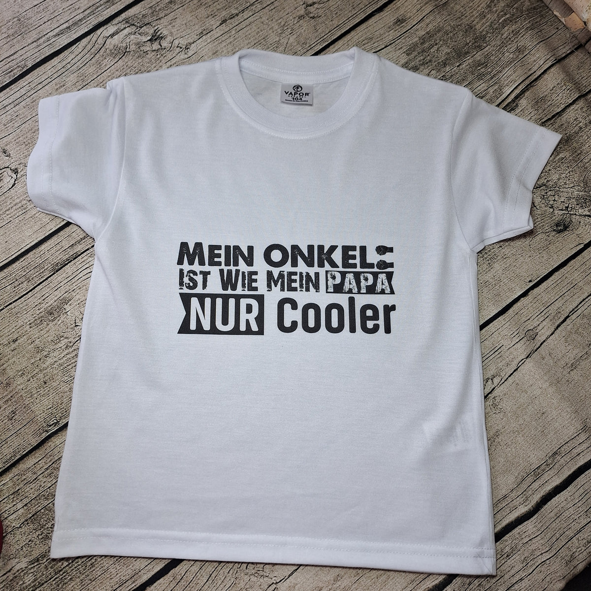 Weißes Kurzarm-Kinder-T-Shirt mit Rundhalsausschnitt und schwarzem Onkel-Spruch, flach auf Holz ausgelegt.