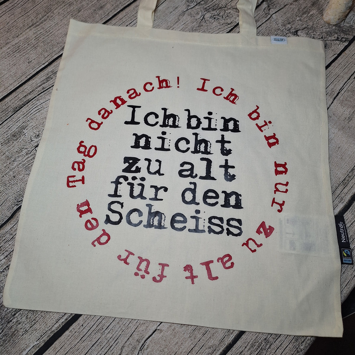 Naturfarbene Stofftasche mit zwei Tragehenkeln und schwarz-rotem Spruchmotiv, flach auf Holz ausgelegt.