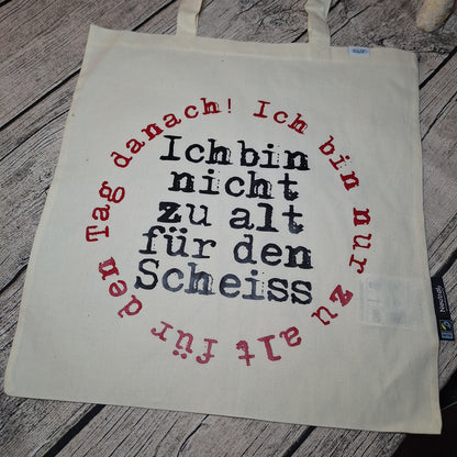 Naturfarbene Stofftasche mit zwei Tragehenkeln und schwarz-rotem Spruchmotiv, flach auf Holz ausgelegt.