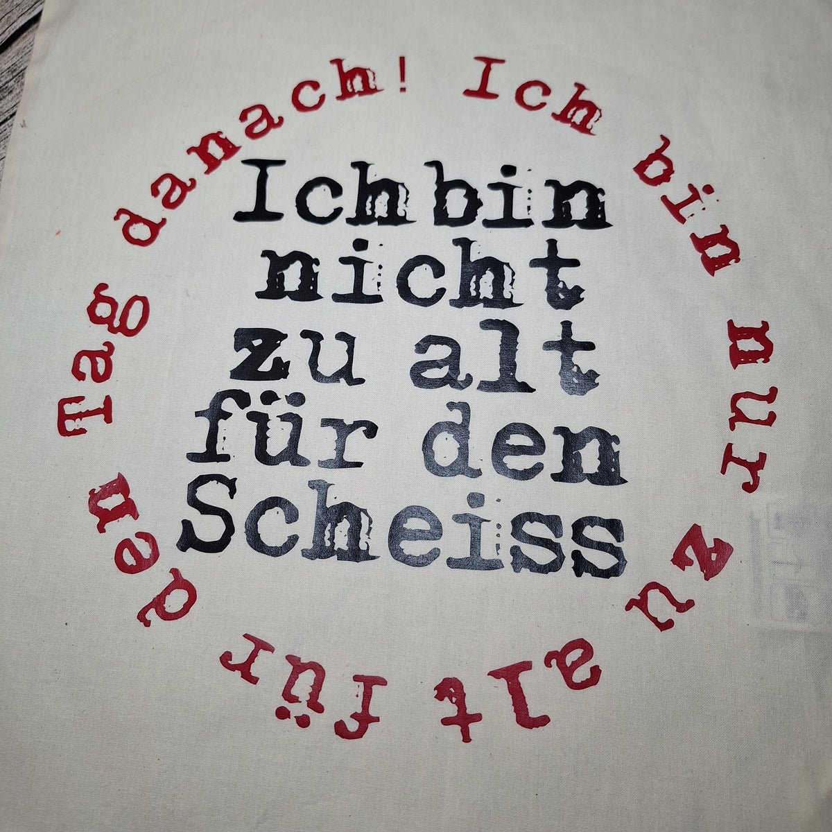 Nahaufnahme des schwarzen Schriftzugs mit kreisförmig angeordnetem rotem Text auf der naturfarbenen Stofftasche.