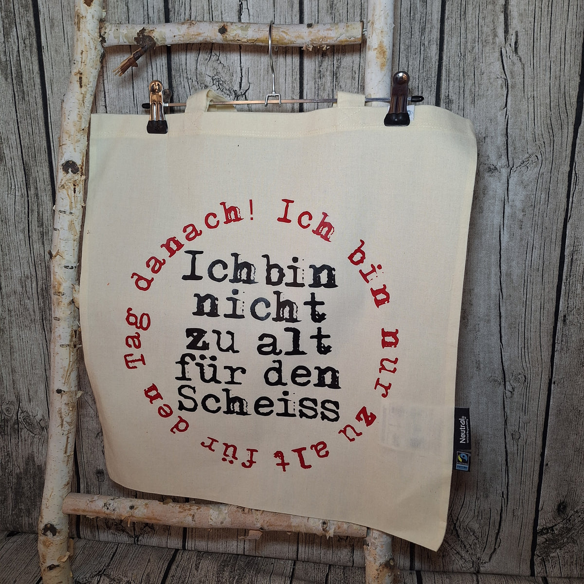 Naturfarbene Baumwolltasche mit schwarzem und rotem Spruch „Ich bin nicht zu alt für den Scheiss. Ich bin nur zu alt für den Tag danach!“, hängend vor Holz.