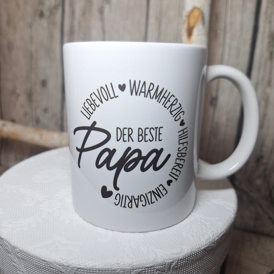 Weiße Keramiktasse mit schwarzem Schriftzug „Der beste Papa“, Herzmotiven und kreisförmig angeordneten Eigenschaften.
