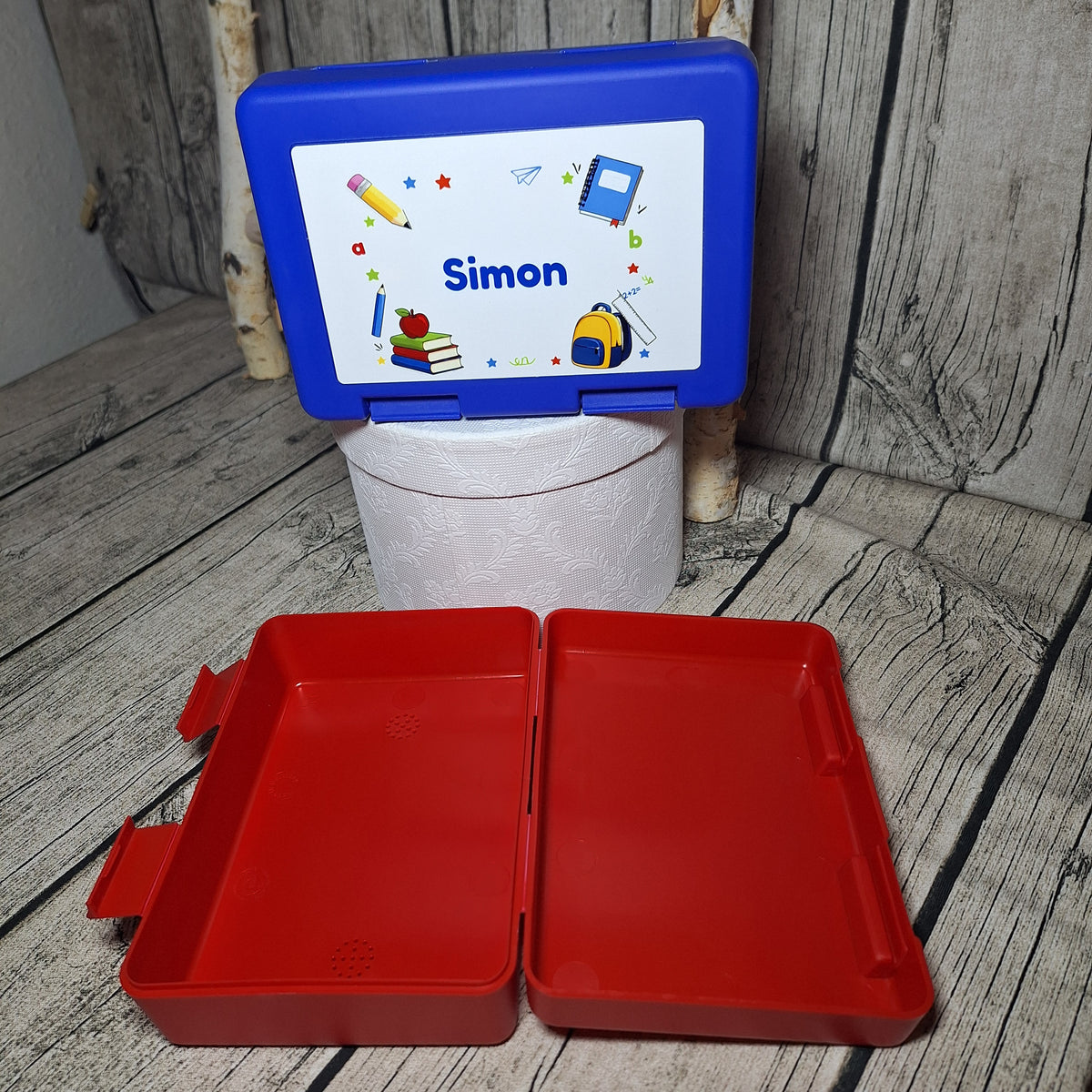 Geöffnete rote Jausenbox mit Blick in den Innenraum, dahinter eine blaue Box mit Namen Simon und Schulmotiven.