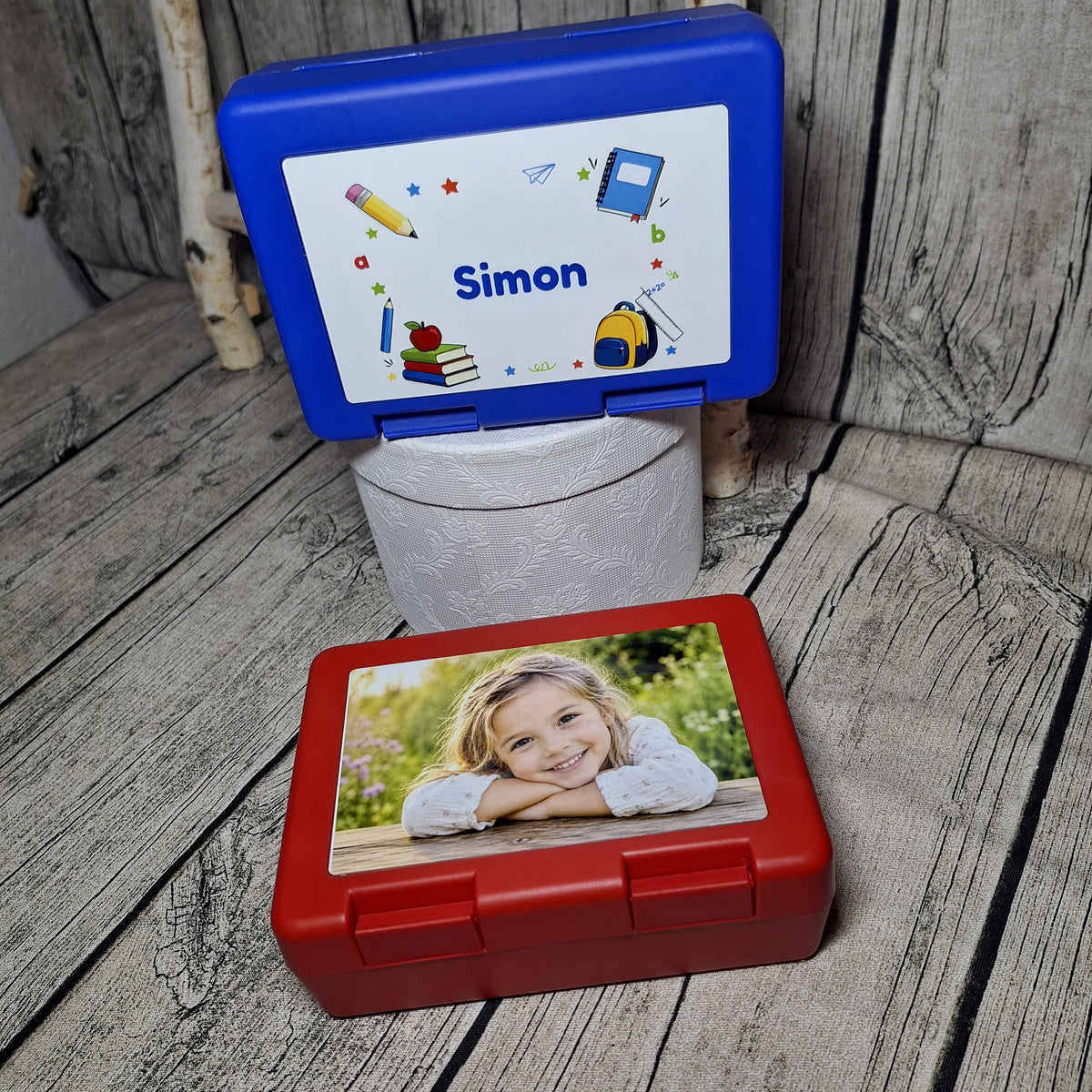 Zwei personalisierte Jausenboxen: blaue Box mit Namen Simon und Schulmotiven sowie rote Box mit Kinderfoto.