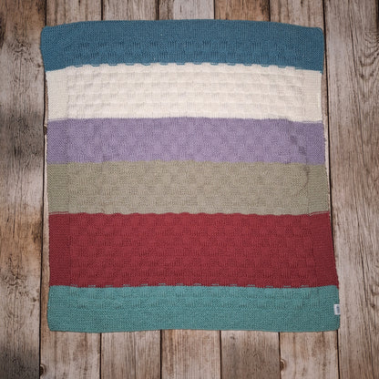 Handgestrickte Babydecke mit breiten Streifen in Petrol, Cremeweiß, Flieder, Grau und Beerenrot auf Holzuntergrund.