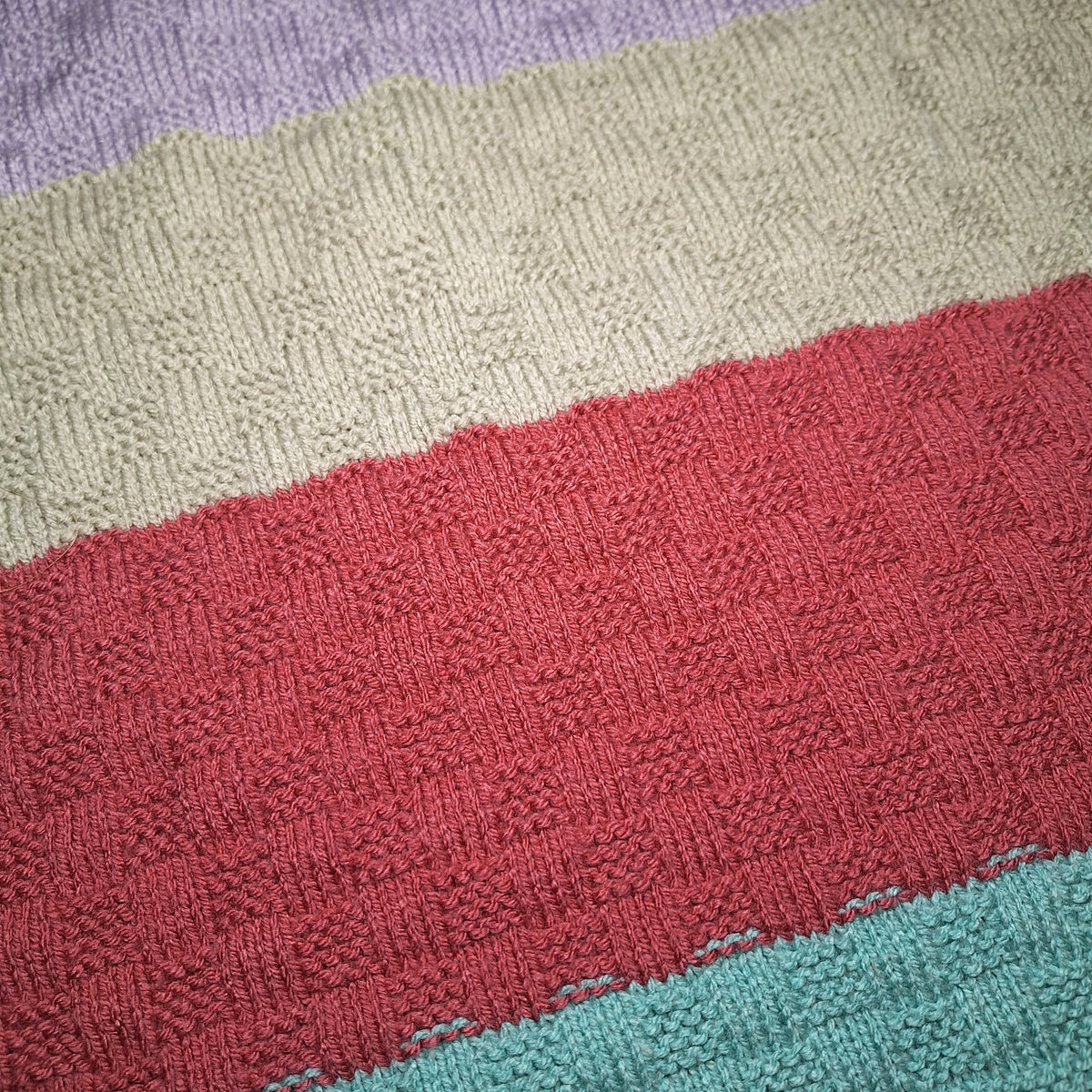 Nahaufnahme der Babydecke mit strukturiertem Strickmuster in Flieder, Grau, Beerenrot und Türkis.