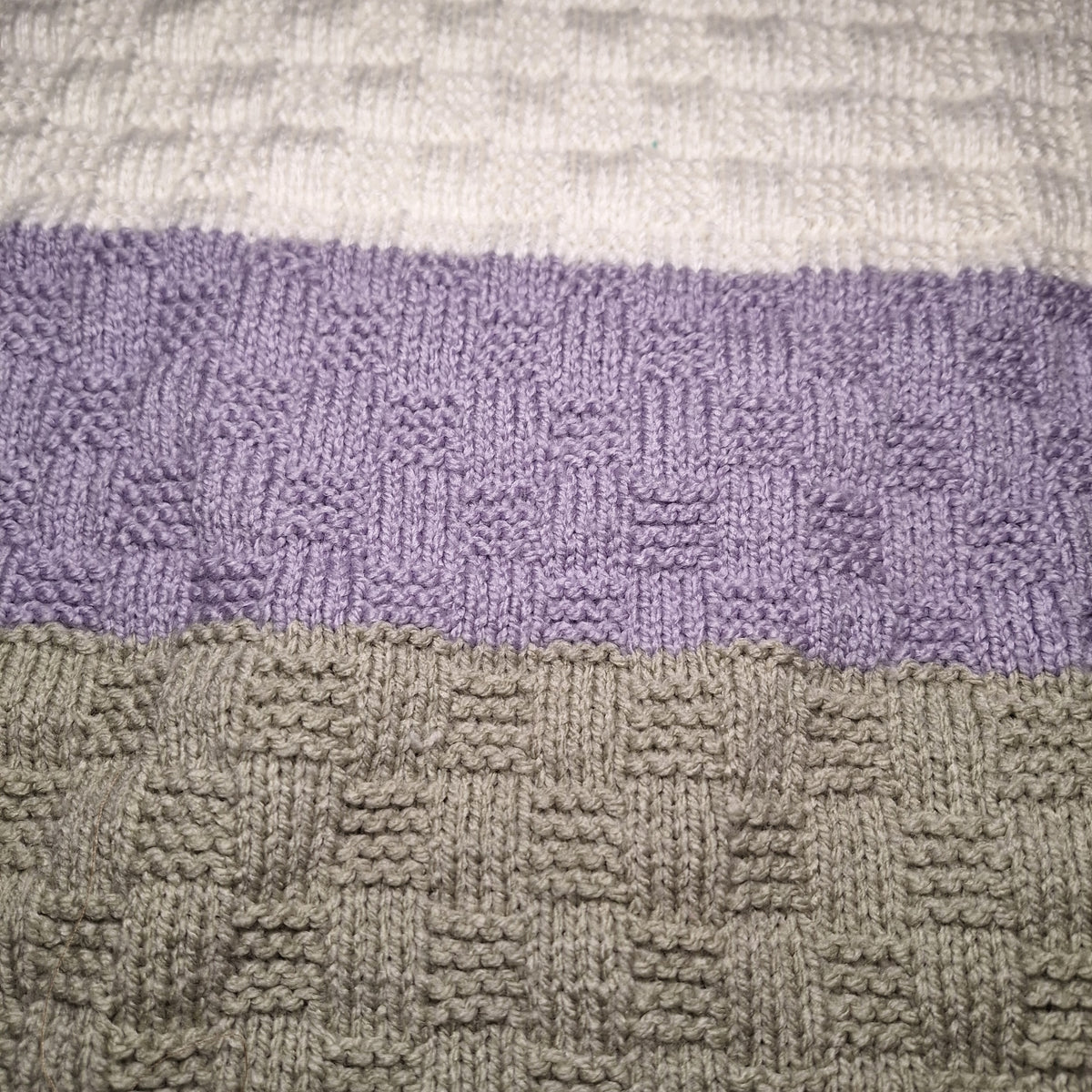 Detail des abwechslungsreichen Strickmusters der Babydecke mit cremeweißen, fliederfarbenen und grauen Streifen.