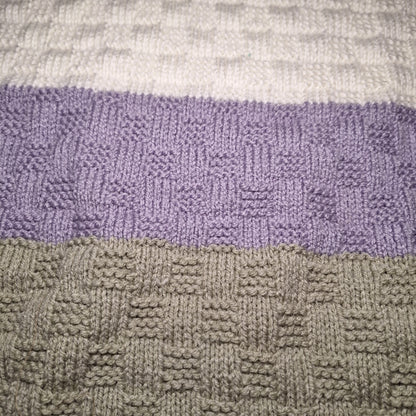 Detail des abwechslungsreichen Strickmusters der Babydecke mit cremeweißen, fliederfarbenen und grauen Streifen.