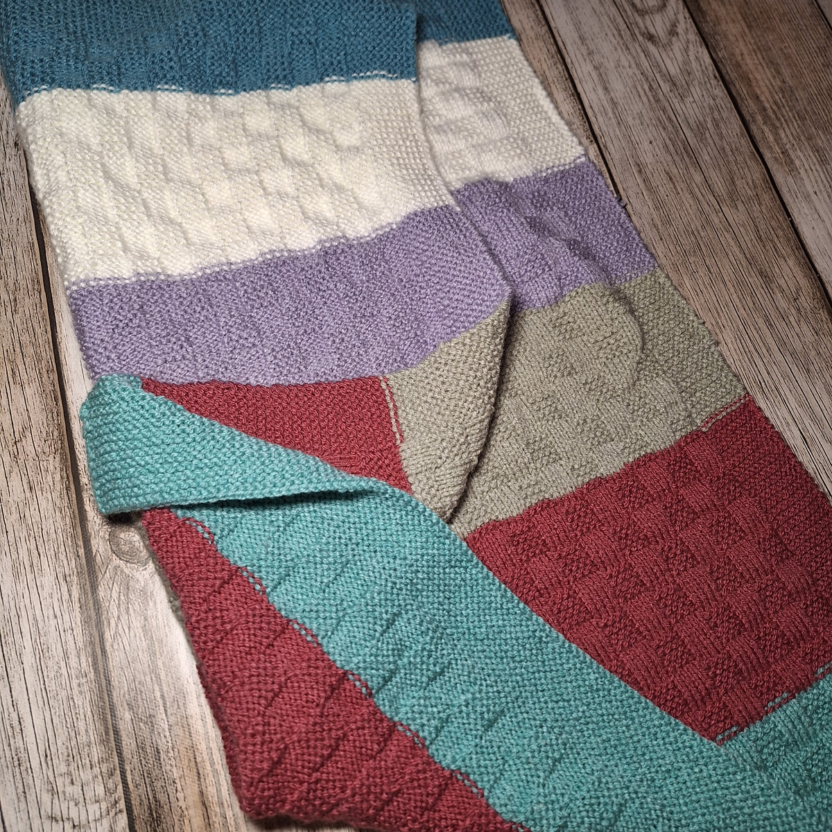 Teilweise umgeschlagene Babydecke mit bunten Streifen, sichtbarer Rückseite und strukturiertem Strickmuster.