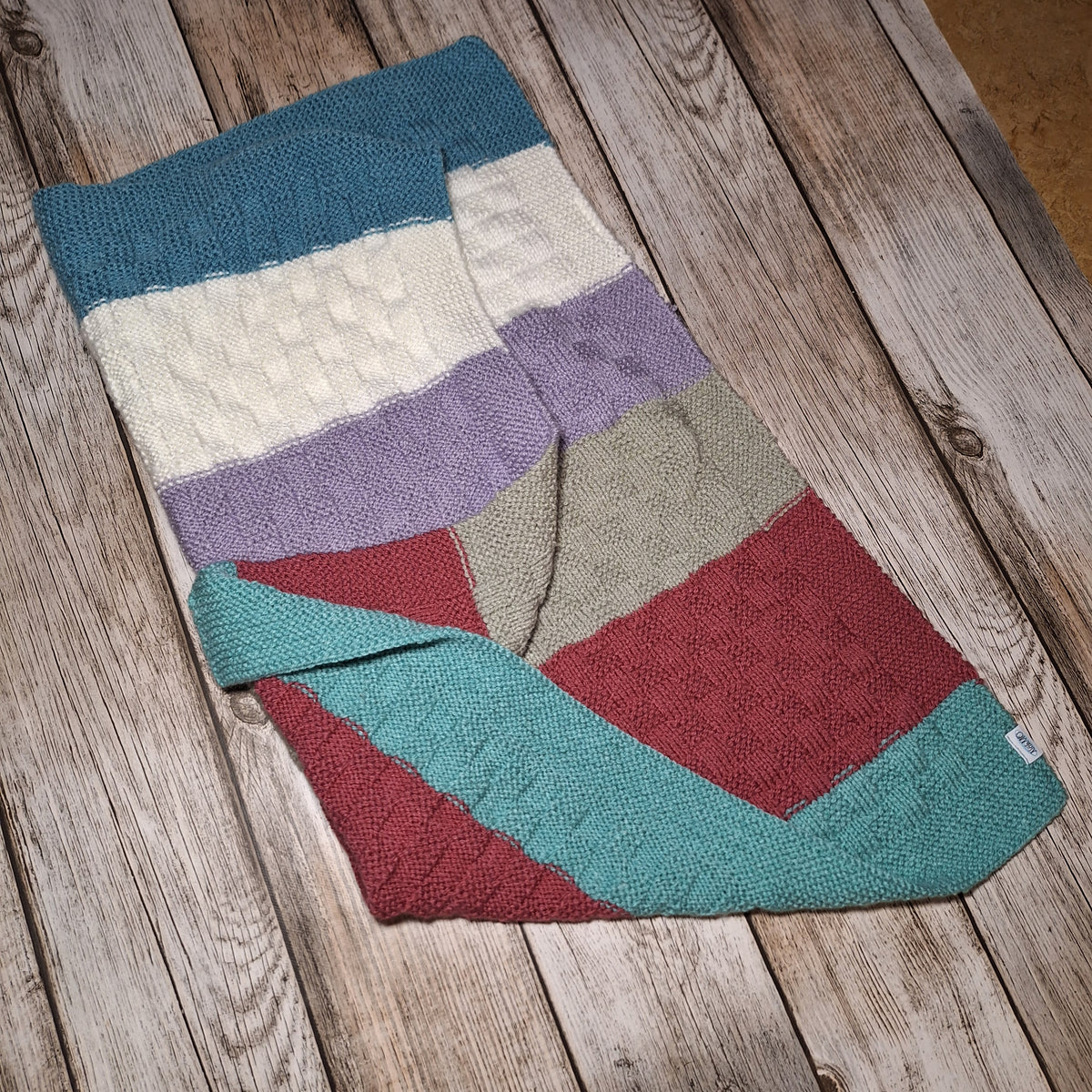 Handgestrickte Babydecke mit bunten Blockstreifen und umgeschlagener Ecke, schräg auf Holzuntergrund fotografiert.