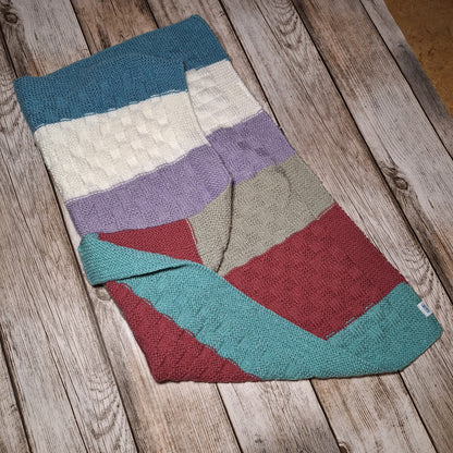 Handgestrickte Babydecke mit bunten Blockstreifen und umgeschlagener Ecke, schräg auf Holzuntergrund fotografiert.