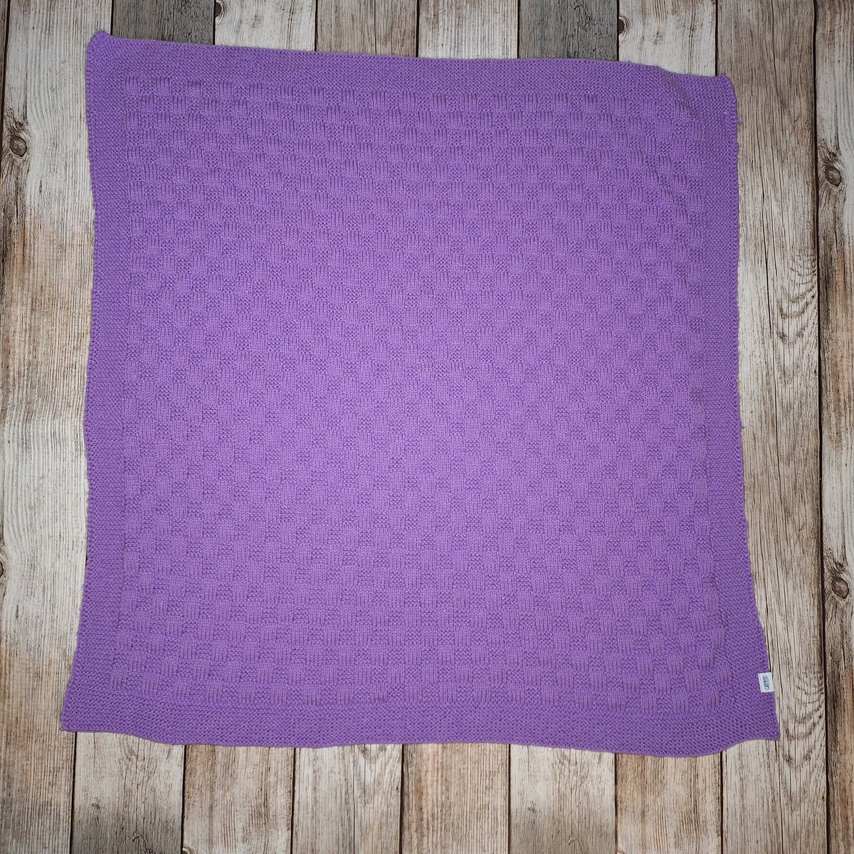 Fliederfarbene handgestrickte Babydecke mit rechteckigem Strukturmuster und umlaufender Bordüre auf Holzuntergrund.