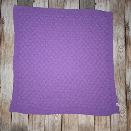 Fliederfarbene handgestrickte Babydecke mit rechteckigem Strukturmuster und umlaufender Bordüre auf Holzuntergrund.