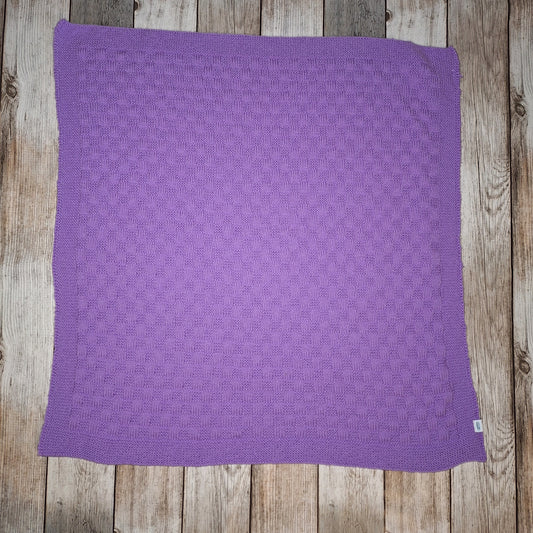 Fliederfarbene handgestrickte Babydecke mit rechteckigem Strukturmuster und umlaufender Bordüre auf Holzuntergrund.