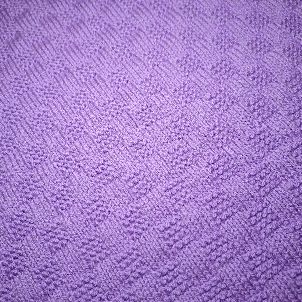 Nahaufnahme des rechteckigen Strukturmusters der handgestrickten Babydecke in Flieder.