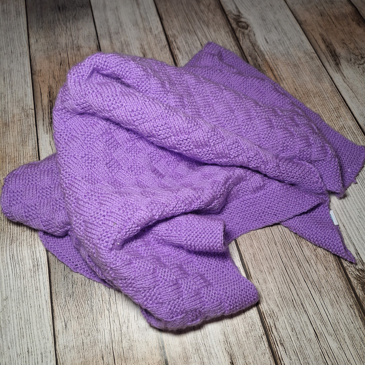 Locker in Falten gelegte fliederfarbene Strickdecke mit sichtbarem Strukturmuster und gestricktem Rand.