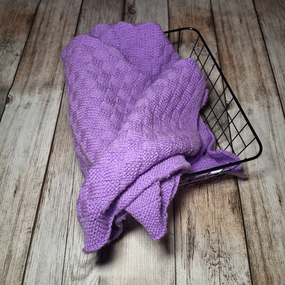 Fliederfarbene handgestrickte Babydecke locker in einem schwarzen Drahtkorb auf Holzuntergrund.