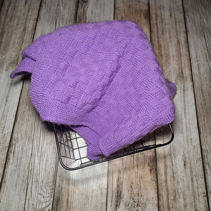 Ansicht von oben auf eine fliederfarbene Strickdecke mit Strukturmuster in einem schwarzen Drahtkorb.