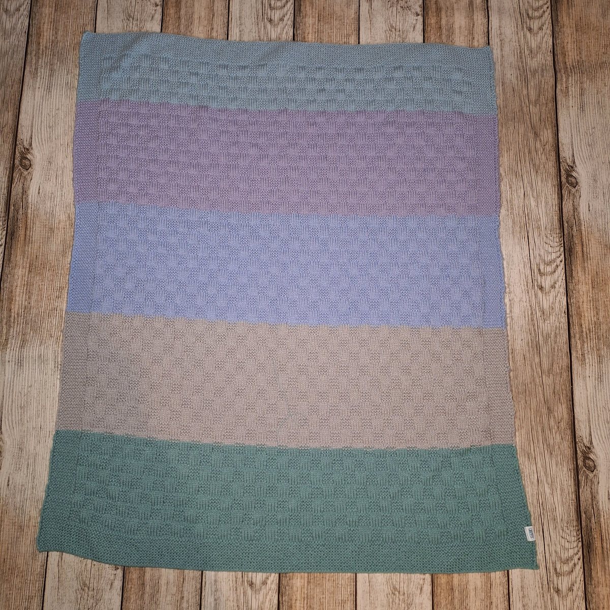 Handgestrickte Babydecke mit breiten Pastellstreifen und rechteckigem Strukturmuster, ausgebreitet auf Holzuntergrund.