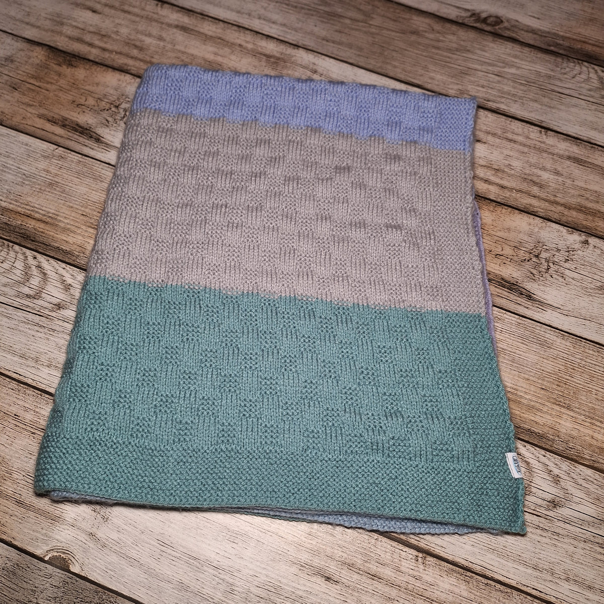 Zusammengelegte gestreifte Babydecke mit Strukturmuster und kleinem Markenetikett auf Holzuntergrund.