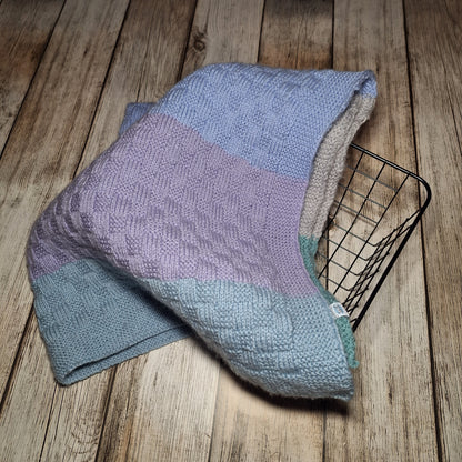 Handgestrickte Babydecke mit Pastellstreifen, locker über einen schwarzen Drahtkorb gelegt.